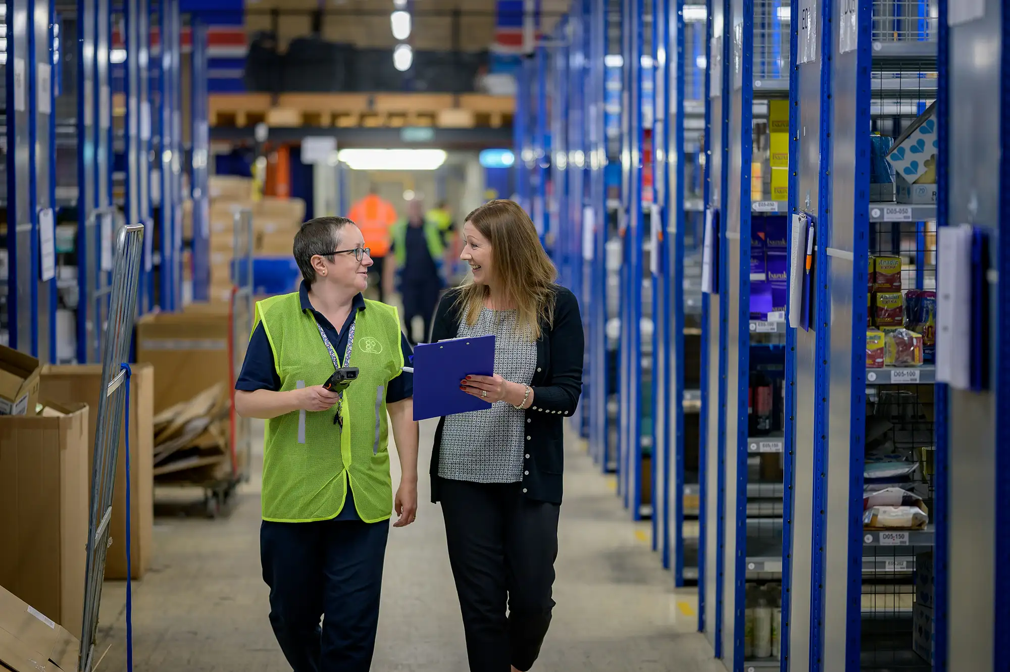 Two people walking through a warehouse aisle with shelving, holding a clipboard and discussing operational checks.